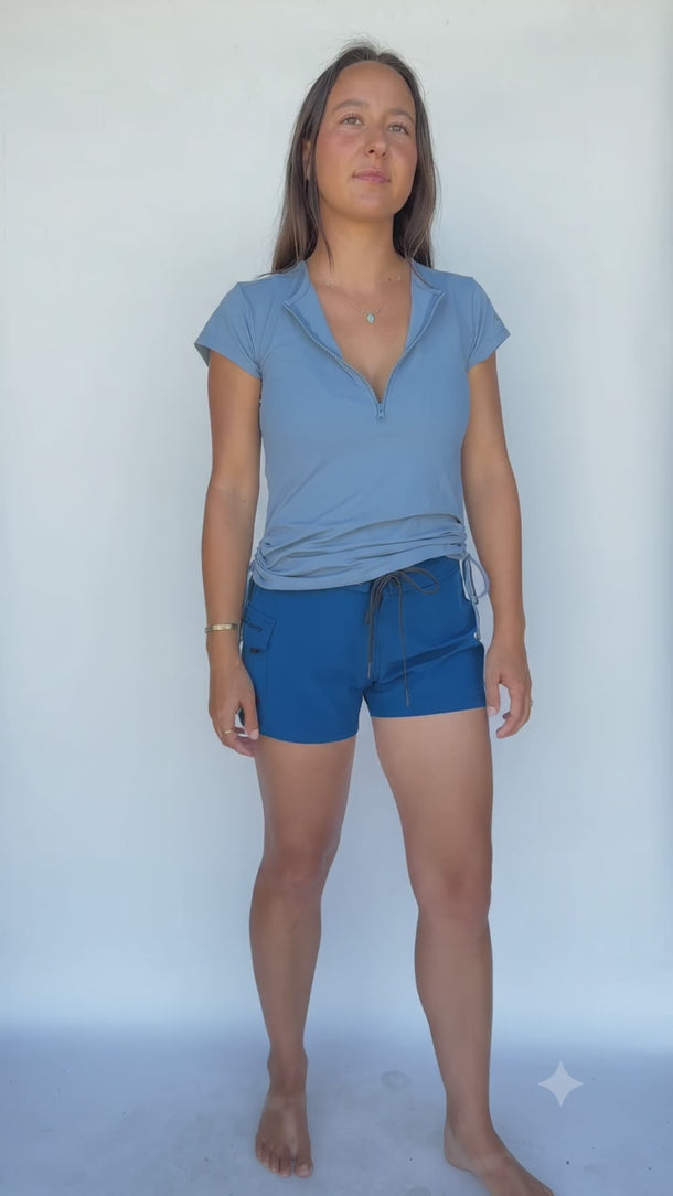 woman turning around showing all angles of her swim top in glacier blue which has strings on each side that cinch the sides up as well as a 3/4 zipper in the bust. She is also wearing the vela blue 3 inch inseam swim shorts.